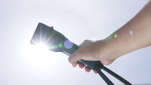 Closeup hand grasping an EV plug for electric vehicle with the midday sky in the background as progressive idea of alternative sustainable clean and green energy for environmental concern.