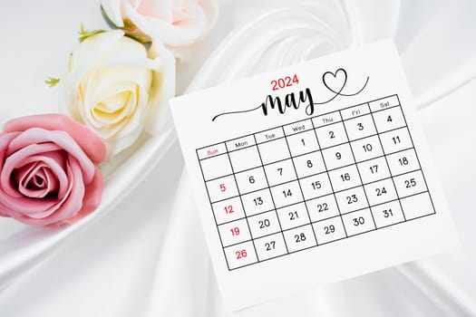 May 2024 calendar page and rose flower on white satin textile background.