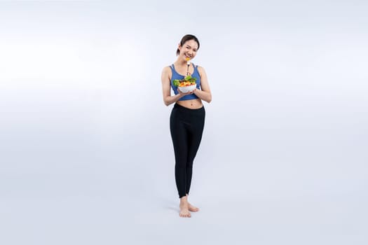 Young sporty Asian woman in sportswear holding salad bowl fill with vibrant of fruit and vegetable. Natural youthful and fit body lifestyle with balance nutrition on isolated background. Vigorous