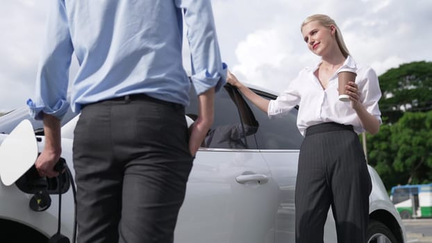 Progressive businessman and businesswoman leaning on electric car connected to charging station before driving around city center. Eco friendly rechargeable car powered by clean energy.