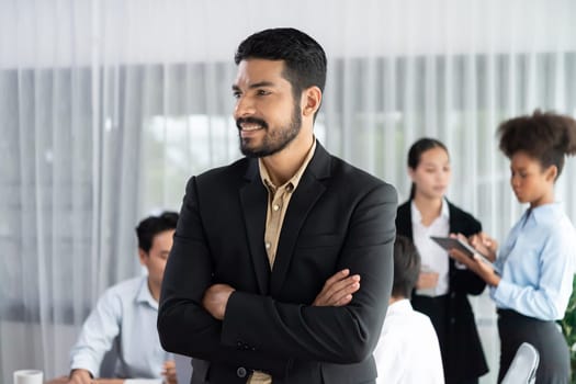 Businessman poses confidently with diverse coworkers in busy meeting room background. Multicultural team works together for business success. Modern businessman portrait. Concord