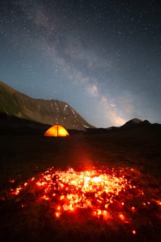 Night landscape long exposure. burning coals are the remains of a fire against the backdrop of a luminous trekking tent of the mountains and the milky way.