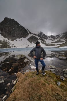 A young man poses and smiles at high altitude after a hike. A male tourist looks to the side against the backdrop of the Caucasus mountains and a lake in ice, in foggy weather