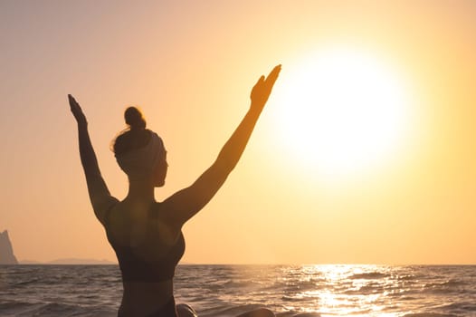 Free happy woman raises her hands and looks at the sun, rear side view. In the background, dawn, greeting the sun during yoga or meditation.