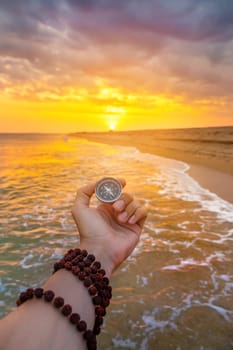 POV of a person holding a navigation compass to find the direction and next destination. Travel and exploration concept. Beach with sandy shore and sea against the backdrop of rolling waves and sunset sky.