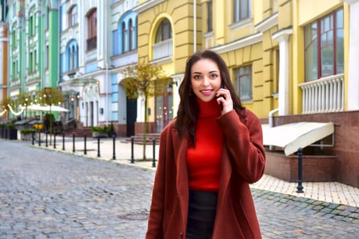 Beautiful stylish lady in a brown coat walking on the street smiles while talking on the phone.