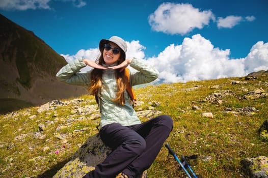 A smiling woman with a backpack travels through the mountains and rests on the top of a mountain on a summer sunny day. Attractive girl enjoying healthy lifestyle outdoors while looking at beautiful nature
