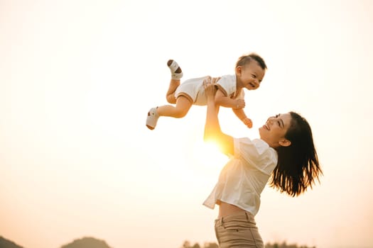 A woman holds her newborn baby up high, as they enjoy a moment of nature in the park at sunset. The little one looks up at the sky with wonder, while the proud mom captures the precious life moment