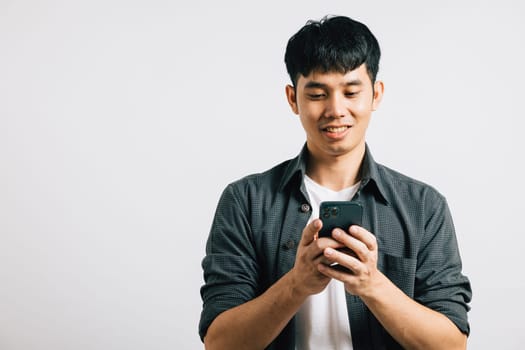 Portrait of a smiling Asian young man, excited while typing a text message on his smart mobile phone for online communication. Studio shot isolated on white, showcasing his enthusiasm for technology.