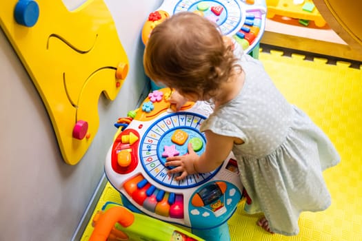 Toddler exploring a colorful activity table in a bright playroom, learning shapes and sounds. Concept of promoting early learning and engaging motor skills development