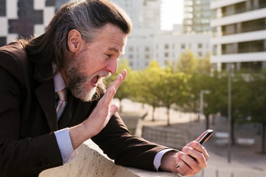 smiling mature man with beard and gray hair in classy suit waving hand while greeting partner during video meeting on smart phone while standing against modern city buildings, copyspace for text