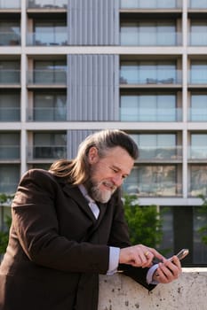 smiling middle age male with beard and gray hair in classy suit browsing on smartphone against modern city buildings, concept of business and mature man lifestyle, copyspace for text