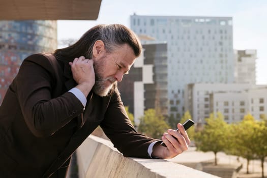 concerned mature male with beard in classy suit using mobile phone while standing against modern city buildings, concept of technology of communication and middle age man lifestyle, copyspace for text