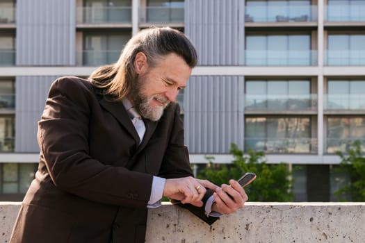 smiling middle age male with beard and gray hair in classy suit browsing on smartphone against modern city buildings, concept of business and mature man lifestyle, copyspace for text