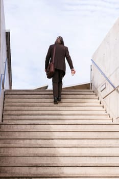 rear view of unrecognizable stylish mature businessman in classy suit with briefcase and takeaway coffee walking up urban stairs in city, concept of business and middle age man lifestyle