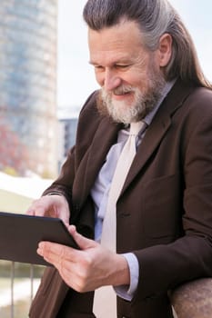 positive middle age male with beard in formal outfit browsing tablet while standing at city, concept of business and mature man lifestyle