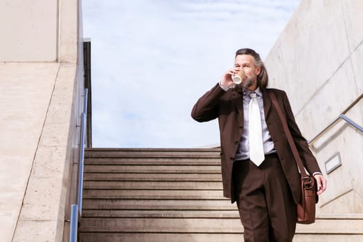 confident mature male executive manager in classy suit with bag drinking takeaway coffee while walking down urban stairs in city, concept of business and middle age man lifestyle, copyspace for text