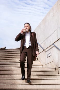 stylish mature businessman in classy suit with briefcase and takeaway coffee walking down urban stairs in city, concept of business and middle age man lifestyle