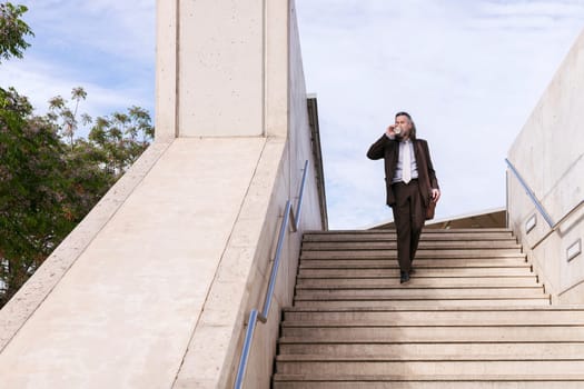 stylish mature businessman in classy suit with briefcase and takeaway coffee walking down urban stairs in city, concept of business and middle age man lifestyle, copyspace for text