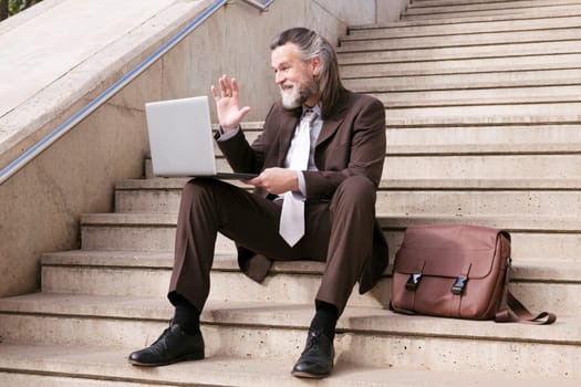 positive middle age male entrepreneur in elegant suit siting on stairway and waving hand saying hello while having online video meeting via laptop, concept of business and middle age man lifestyle