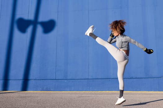 latin female fighter in sportswear and boxing gloves jumping with raised leg while exercising on blue background, concept of strength and healthy lifestyle, copyspace for text