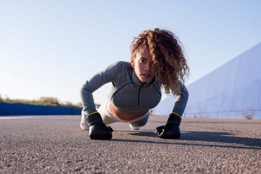 latin sports woman in sportswear doing push ups with boxing gloves on asphalt pavement, concept of urban sport and healthy lifestyle