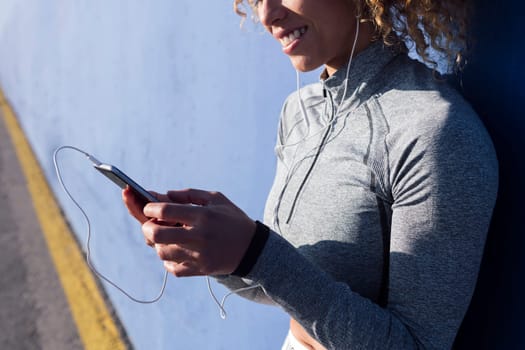 content female athlete in earphones surfing internet on cellphone during break from workout on blue background, concept of urban sport and technology of communication