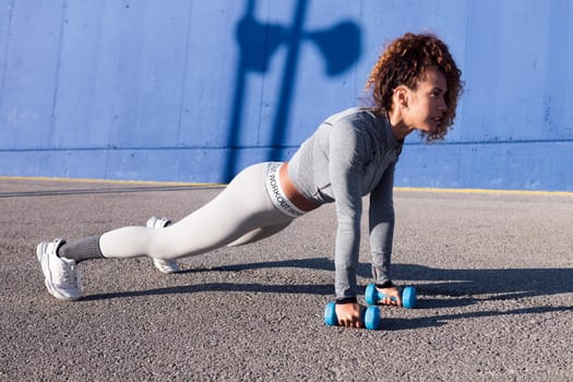 fit female athlete in plank pose with dumbbells and looking forward during training on pavement, concept of urban sport and healthy lifestyle