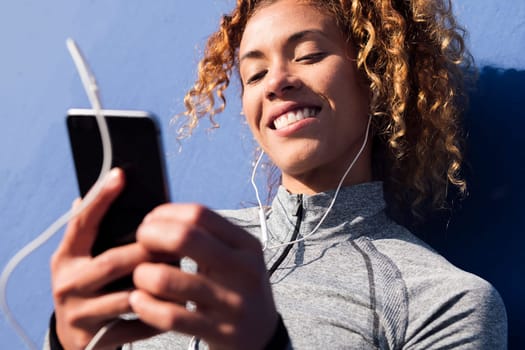 latin female athlete in earphones smiling happy while using mobile phone during break from workout on blue background, concept of urban sport and technology of communication
