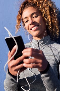 latin female athlete in earphones smiling happy while using mobile phone during break from workout on blue background, concept of urban sport and technology of communication