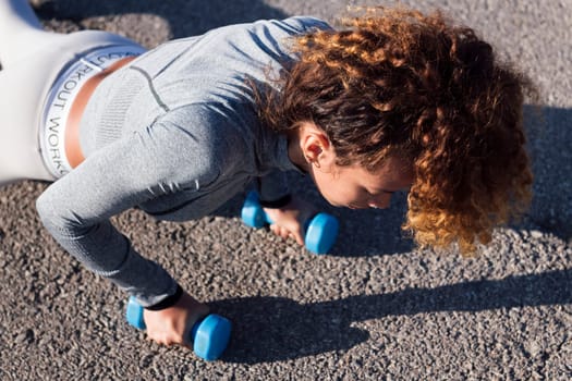 strong female athlete doing planks with dumbbells during training on pavement, concept of urban sport and healthy lifestyle