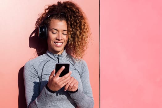 cheerful sportswoman with cellphone listening to music from headset against pink wall, concept of technology of communication and urban lifestyle