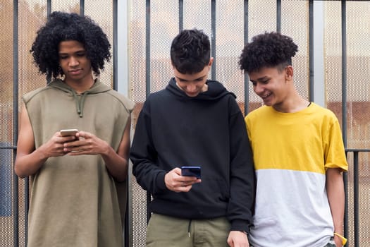 three multiracial teen friends browsing Internet on mobile phones standing against metal fence and looking at screen, concept of youth and technology of communication