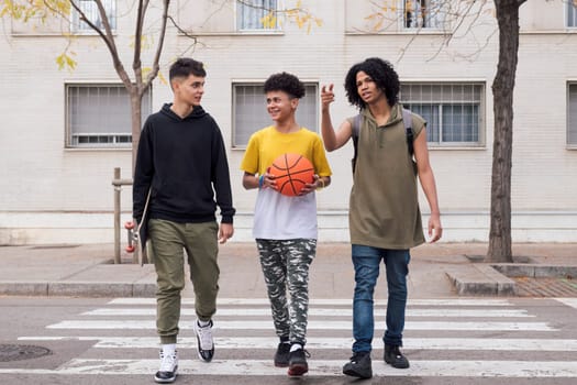 three diverse teen friends with basketball and skateboard talking while crossing road on crosswalk, concept of youth and urban lifestyle