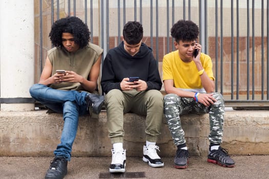 three serious multiracial teen friends browsing Internet on mobile phones sitting against metal fence and looking at screen, concept of youth and technology of communication