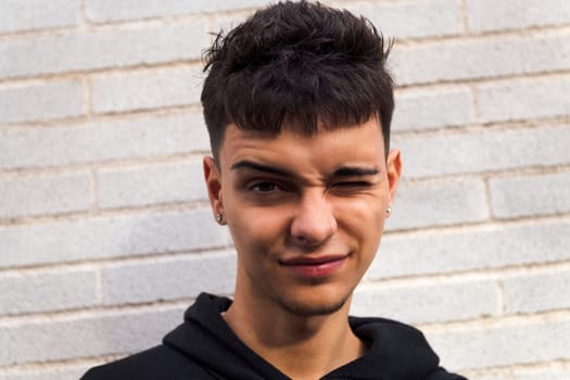 close up of a caucasian male teen wearing casual clothes standing in sunlight against white brick wall winking looking at camera, concept of youth and urban lifestyle, copy space for text