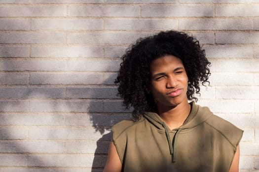 confident african american male teen with afro hairstyle wearing casual clothes standing in sunlight against white brick wall, concept of youth and urban lifestyle, copy space for text