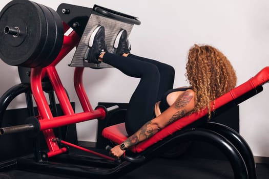 unrecognizable sportswoman with curly hair doing exercises on leg press machine in gym, concept of fitness and active lifestyle