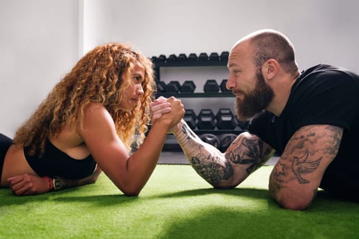 side view of smiling hispanic sportswoman with curly hair holding hand of cheerful bodybuilder during arm wrestling challenge against dumbbells in gym, concept of fitness and active lifestyle