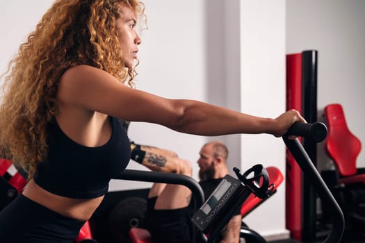 side view of hispanic sportswoman with long curly hair in activewear doing cardio training against bodybuilder in gym, concept of fitness and active lifestyle