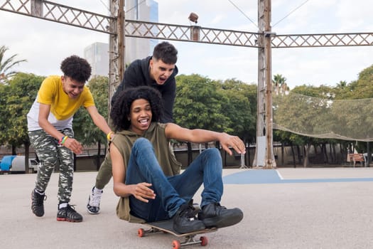 screaming and laughing multiethnic teenagers having fun riding friend on skateboard at sports ground, concept of youth and urban lifestyle
