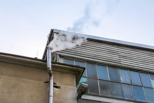 White smoke rises from an exhaust pipe of an industrial building against a clear sky, indicating possible manufacturing activity or heating.