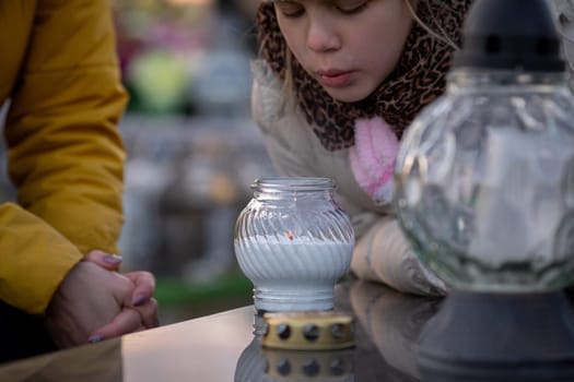 A child is gently blowing on a flickering candle at a gravesite, symbolizing deep remembrance and reflection