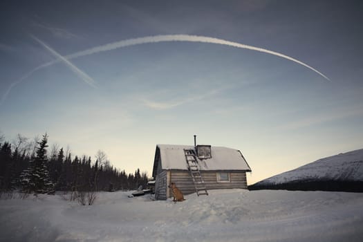 A dog sits and waits outside a hut in the middle of a snowy landscape with a clear sky and a contrail overhead.