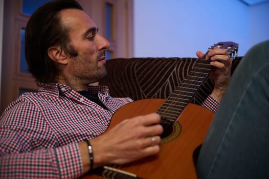 A man sits comfortably on a couch, engrossed in playing his acoustic guitar. He appears relaxed and enjoying the music. The living room ambiance enhances the cozy atmosphere of the scene.
