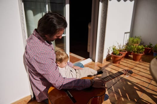 A father and his small child enjoy bonding together outdoors while playing guitar.