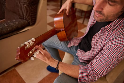 A close-up of a man sitting in a cozy room, playing a wooden stringed instrument with focus and care. The photo highlights the fingering on the fingerboard and represents music and relaxation.