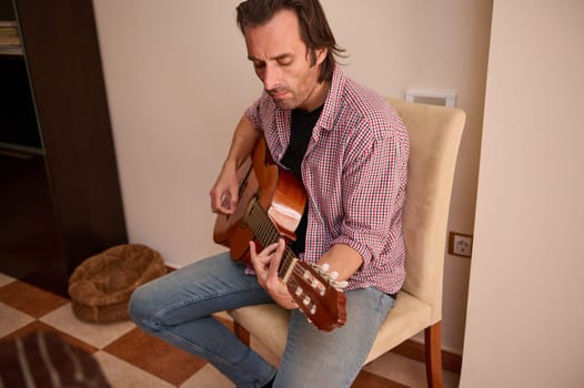 A man in casual clothing is seated indoors, playing an acoustic guitar in an engaging manner, showcasing relaxation and musical inspiration.
