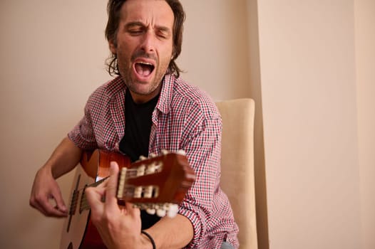 A man in casual attire playing an acoustic guitar and singing passionately indoors. The image conveys emotions and artistic expression, embodying creativity through music in an intimate setting.