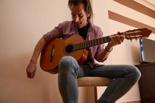 A man in casual attire passionately plays a classical acoustic guitar indoors, concentrating on the melody and tuning the instrument strings, captured during a calm and dedicated moment in music.